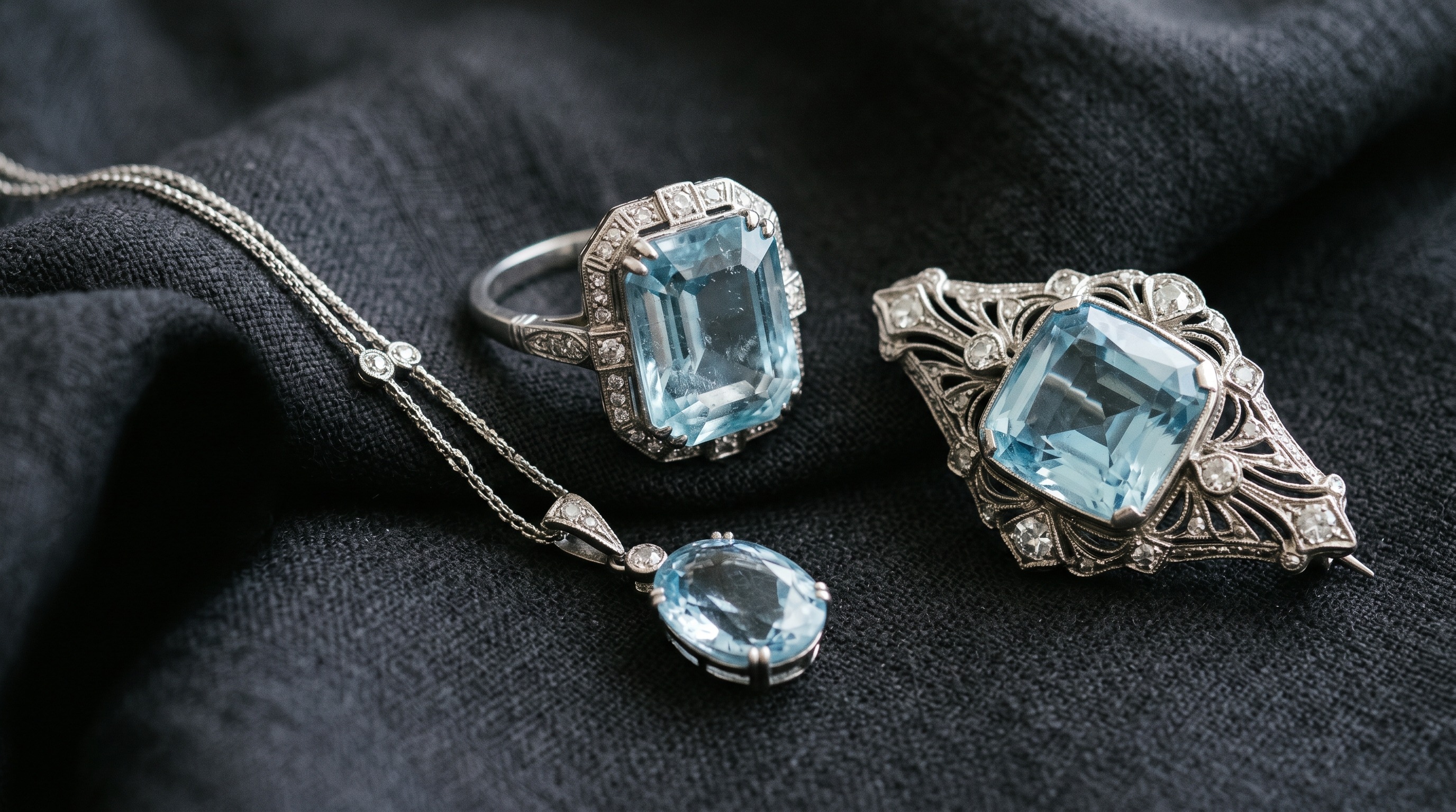 Flat lay of antique aquamarine jewellery including a ring, brooch and pendant on dark fabric