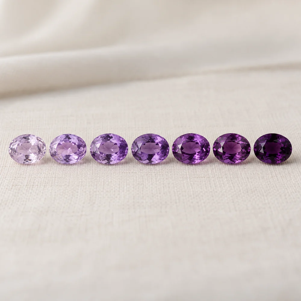 Seven loose oval amethysts in a row, from pale lilac on the left to deep reddish purple and near black purple on the right
