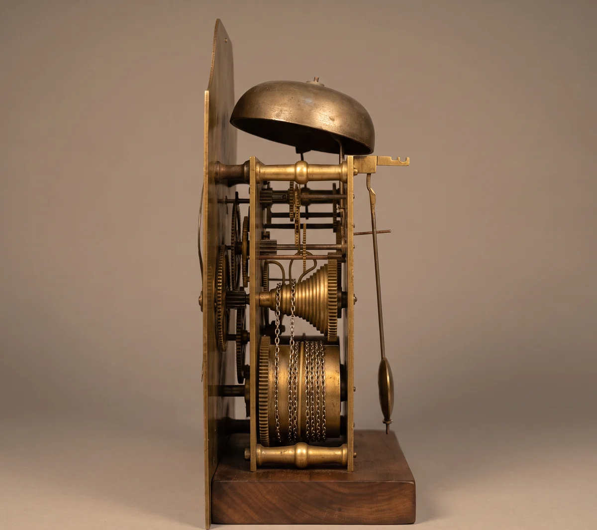 Side view of an English fusee bracket clock movement showing brass plates and pillars, the cone shaped fusee with its chain, the spring barrel, bell and pendulum