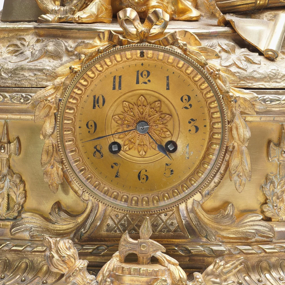Gilt dial of a French mantel clock photographed straight on, with Arabic numerals, a ribbon bow mount above and winding holes either side of the centre