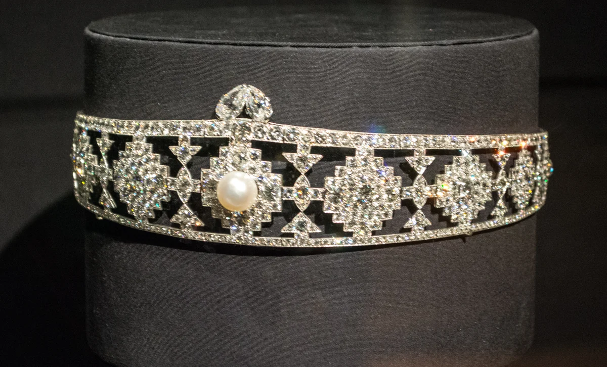 An Art Deco diamond and platinum bandeau by Cartier, designed in 1924: a straight band of geometric openwork diamond panels with a natural pearl at the centre, on a black display form