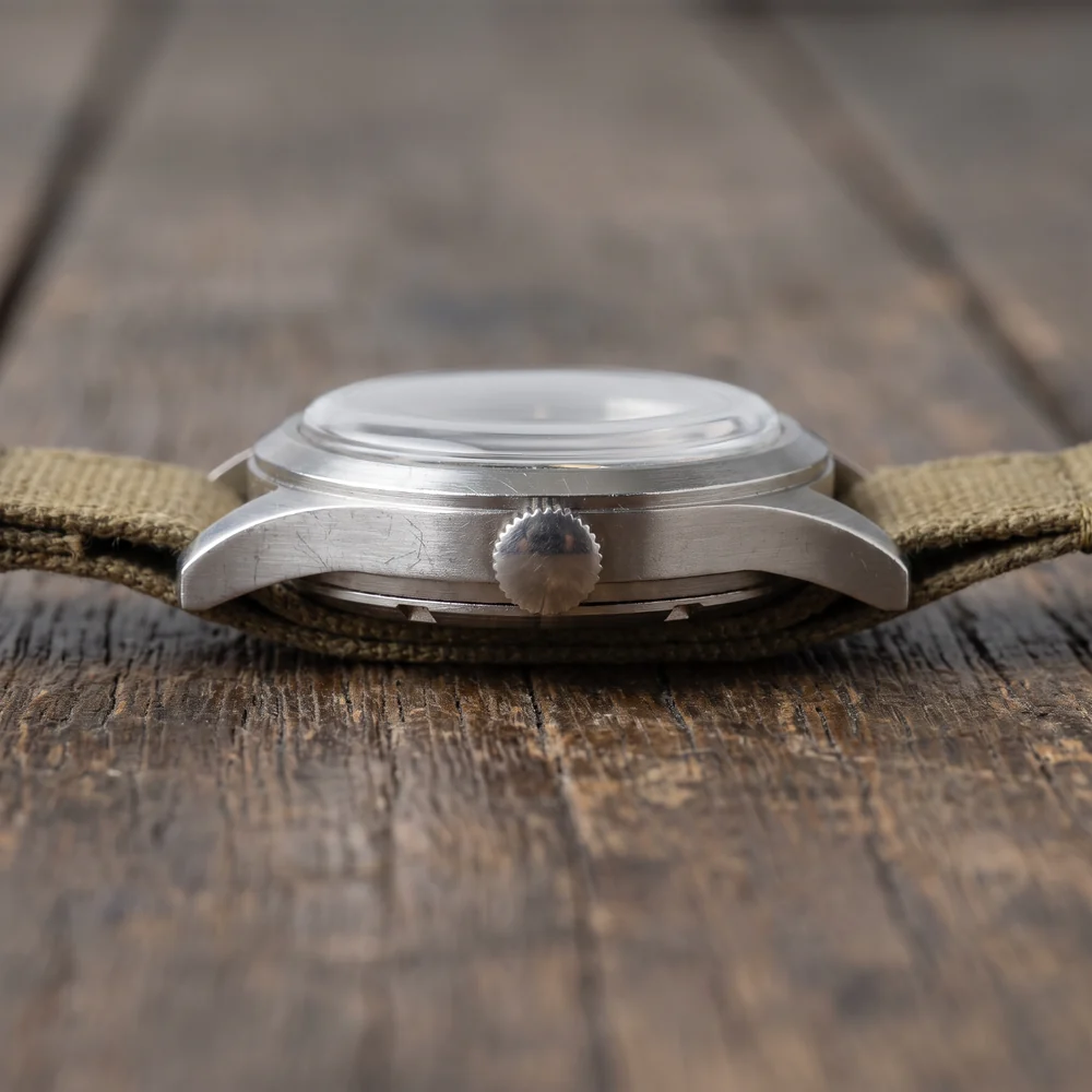 Side view of a brushed steel 1940s military wristwatch on a khaki canvas strap, showing the knurled crown, the lugs and the notched edge of the screw-down case back