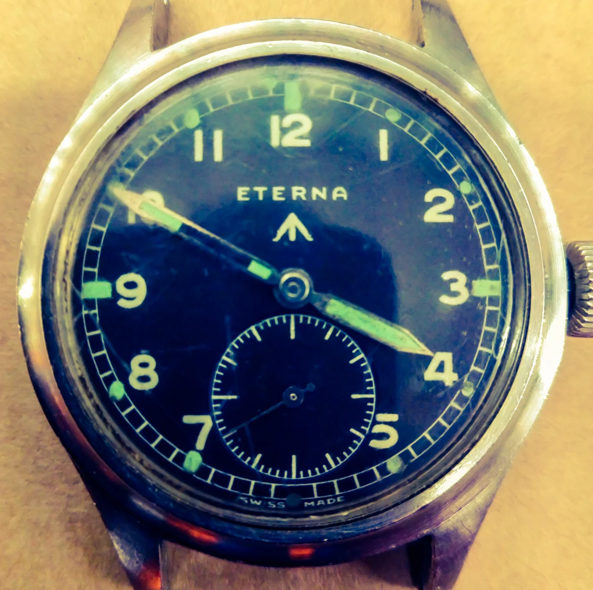 The dial of an Eterna W.W.W. British military wristwatch of 1945: black dial, luminous Arabic numerals and hands, railway minute track, small seconds at six and the broad arrow below the Eterna name