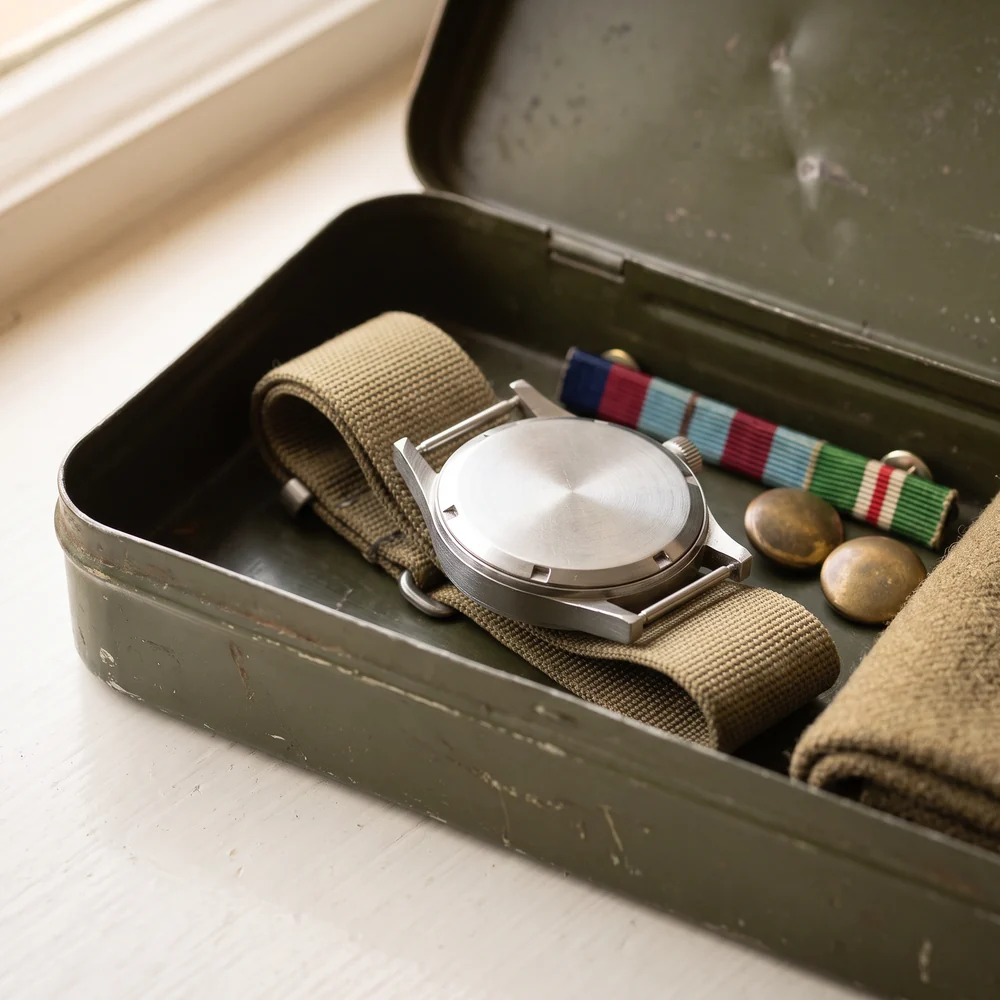 A steel military wristwatch lying face down on its khaki canvas strap in an old green tin, beside a medal ribbon bar and two brass buttons