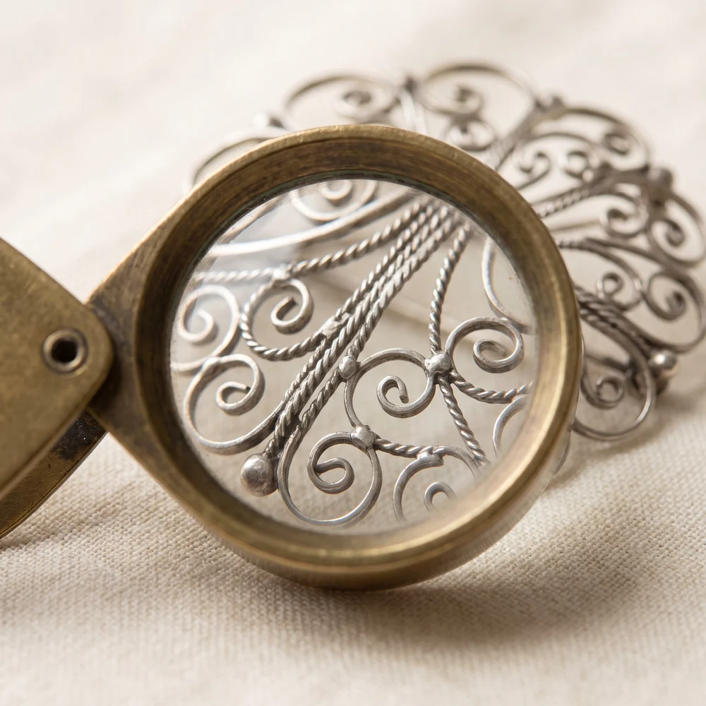 A jeweller's loupe over a silver filigree brooch, magnifying twisted wire, open spirals and small solder joints