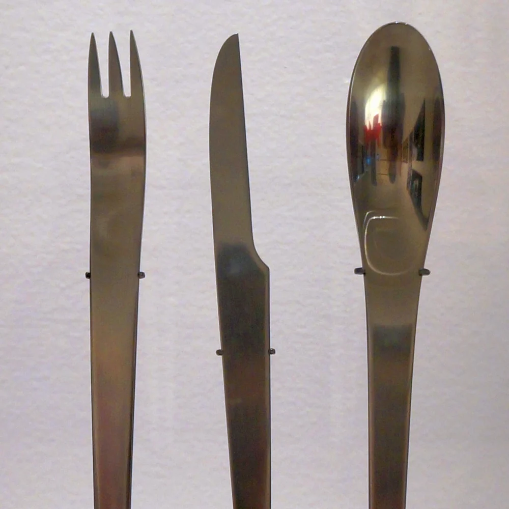 A stainless steel fork, knife and spoon of Arne Jacobsen's 1957 design, standing upright in a display case
