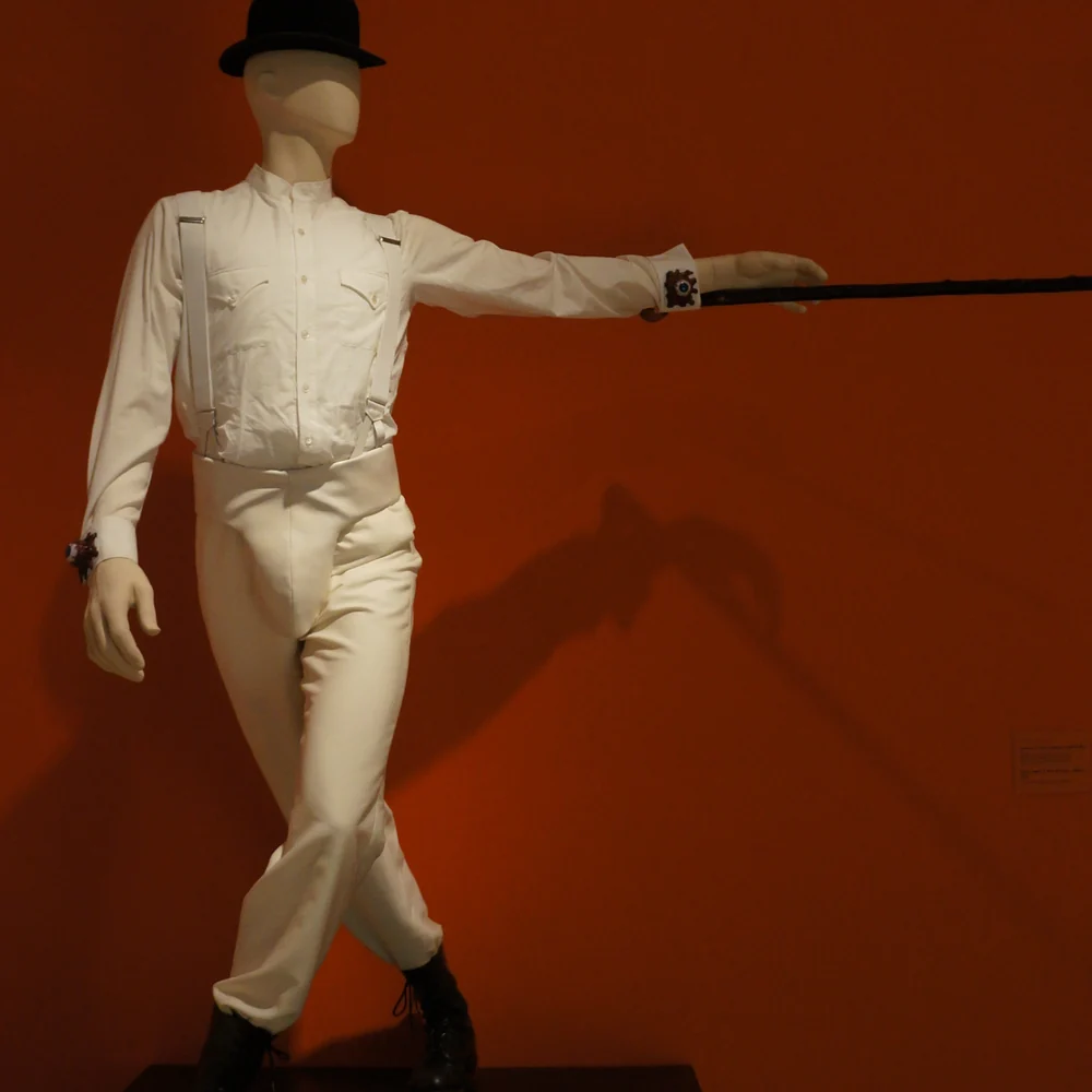 A white shirt and trousers costume with braces, a bowler hat and a cane on a faceless display mannequin against a red wall