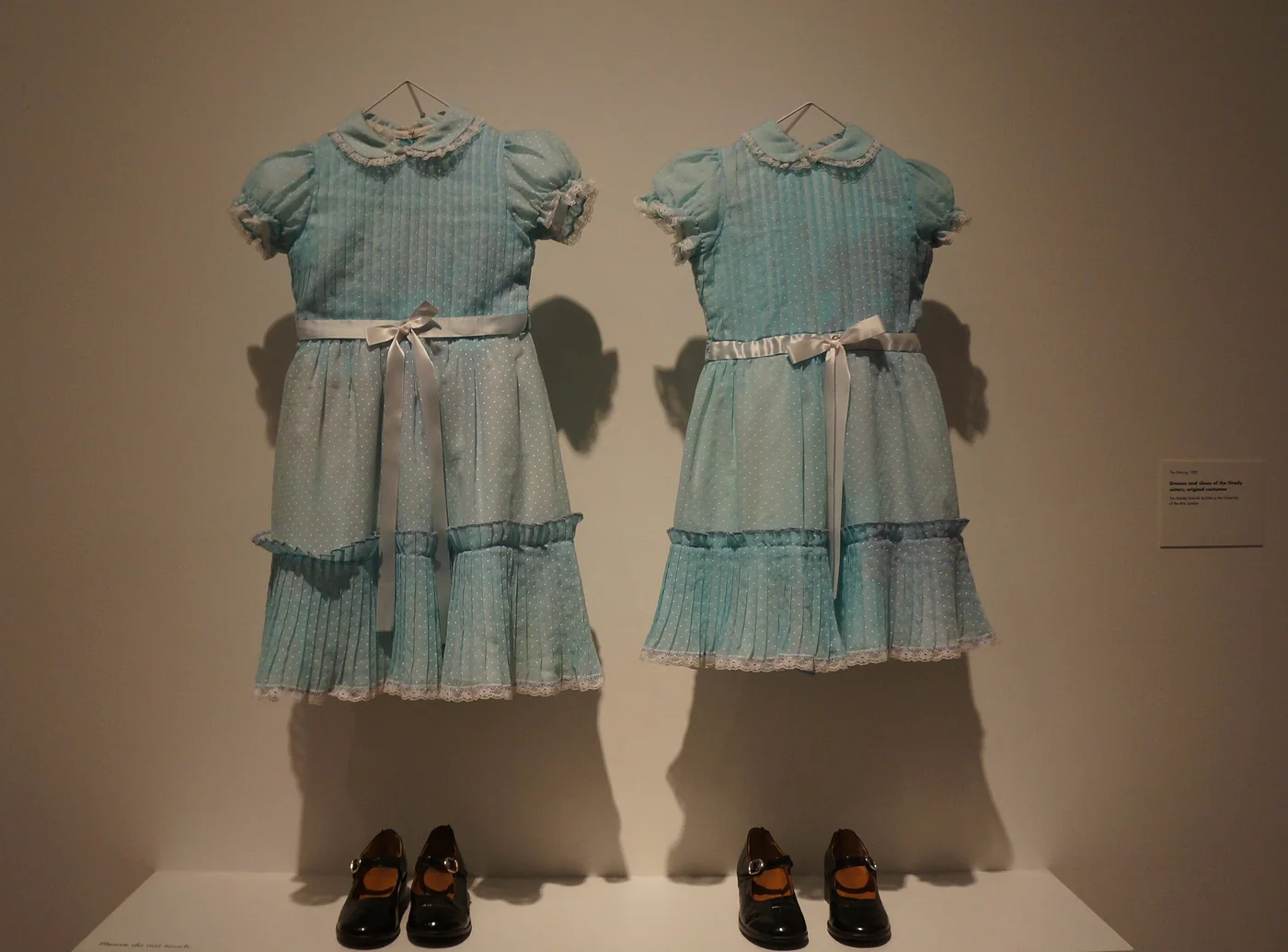 Two pale blue dresses with white collars and sashes, the Grady twins' costumes from The Shining, displayed side by side with two pairs of black shoes