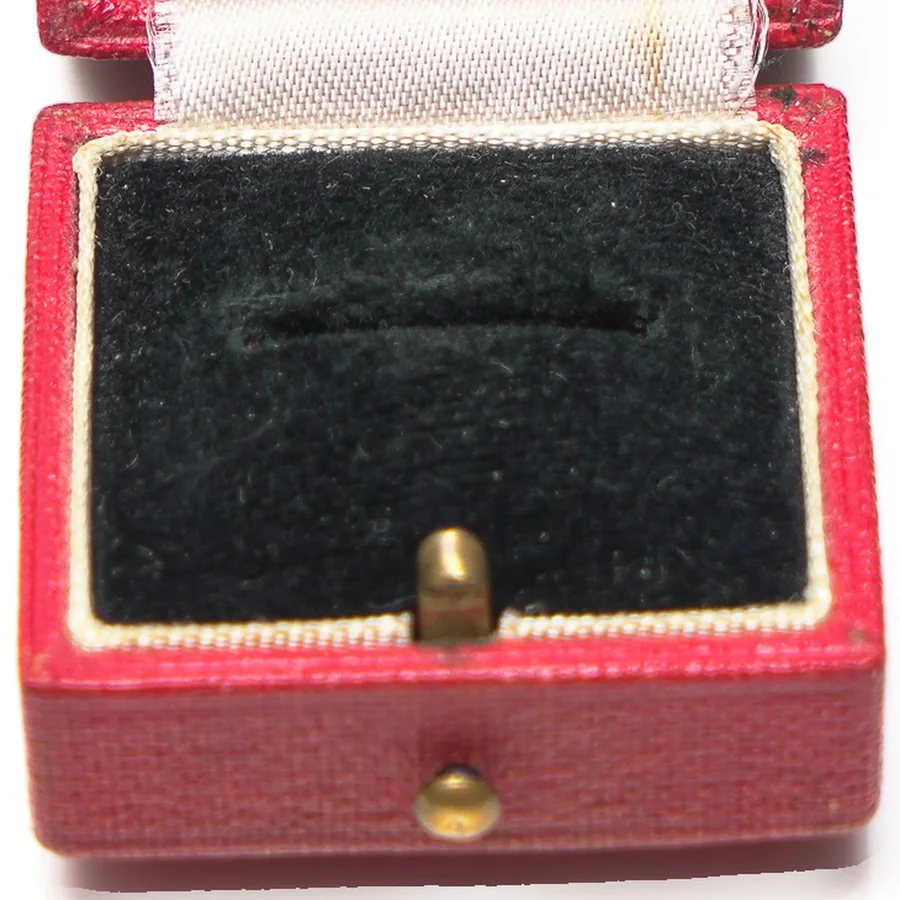 A red leather ring box open to a black velvet pad cut with a single ring slot, with a brass push button at the front