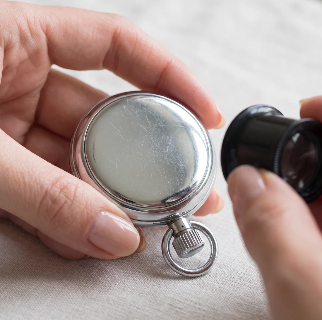 A woman's manicured hands holding a chrome pocket watch face down and checking the case back with a jeweller's loupe