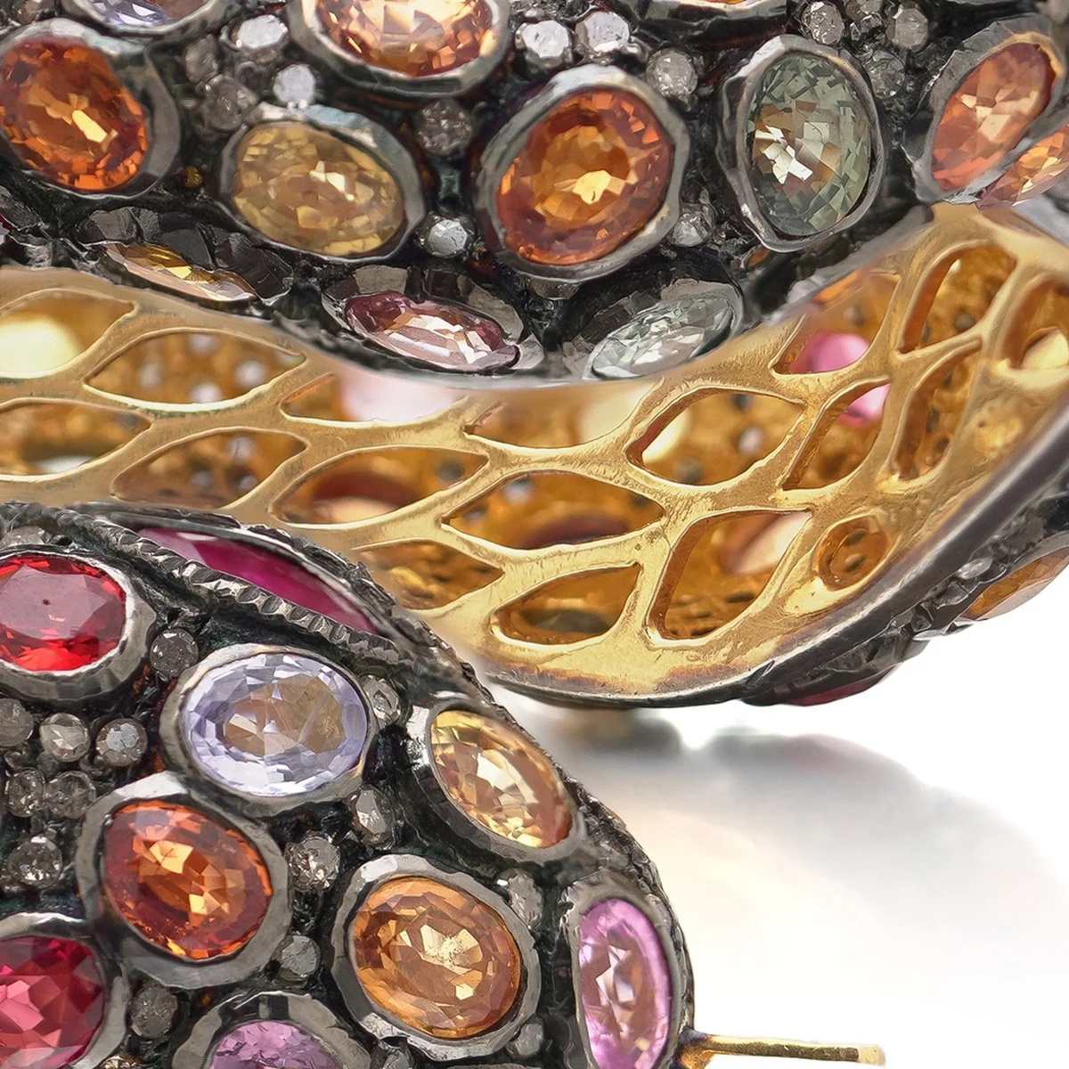 Close-up of multi-coloured sapphires in orange, pink, yellow and violet, close-set in darkened silver with small rose cut diamonds between them, above a pierced gilt lattice