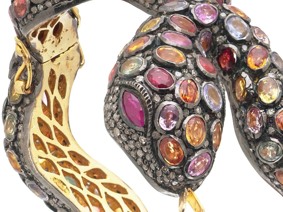 The raised head of a serpent bangle set solid with pink, red and orange sapphires in dark silver, with the gilt honeycomb interior of the body beside it