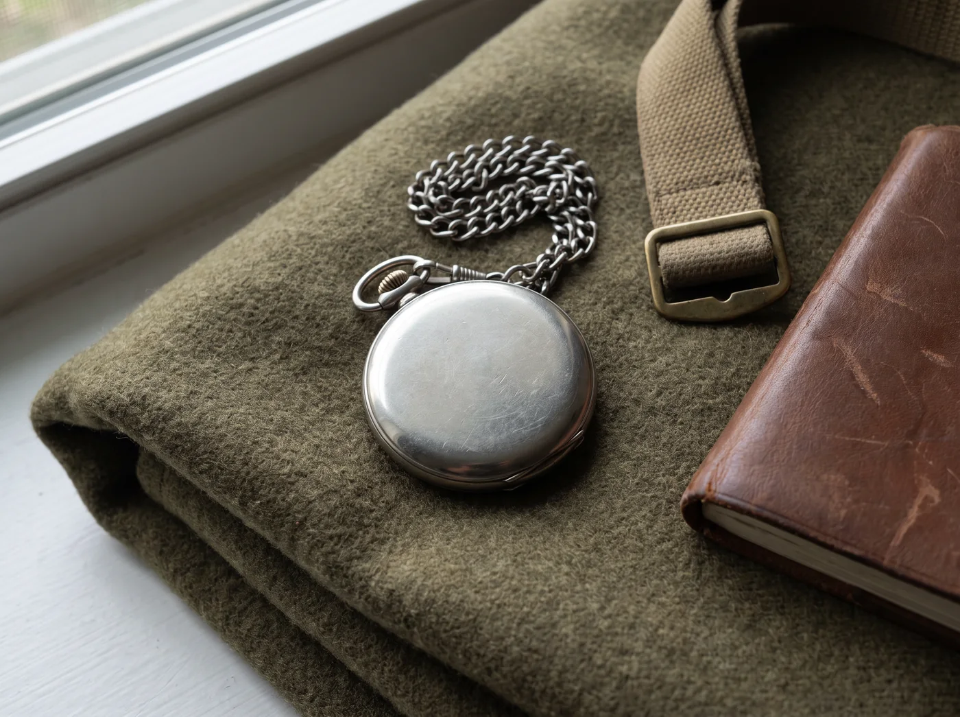 A plain base metal pocket watch lying face down on an olive drab army blanket beside a khaki webbing strap