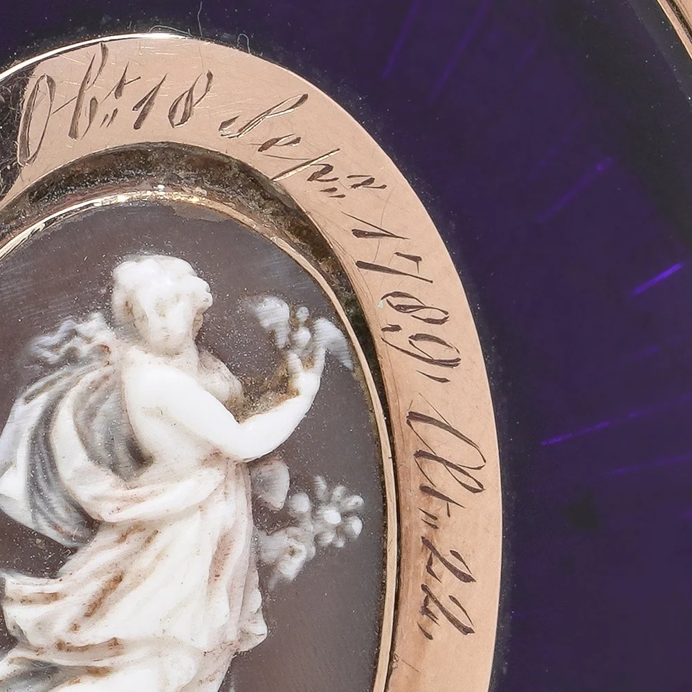 Close-up of the engraved gold frame of a Georgian mourning pendant reading Ob 18 Sepr 1789 Aet 22 beside a carved cameo of a classical woman holding flowers, on deep purple enamel