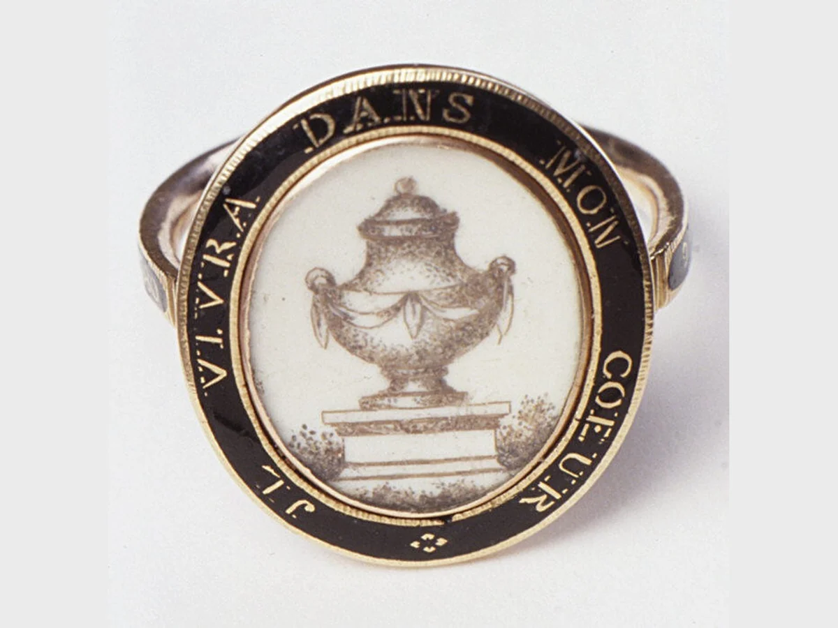 A Georgian mourning ring of about 1780: an oval bezel painted in sepia with a draped urn on a plinth, framed by a black enamel border lettered in gold