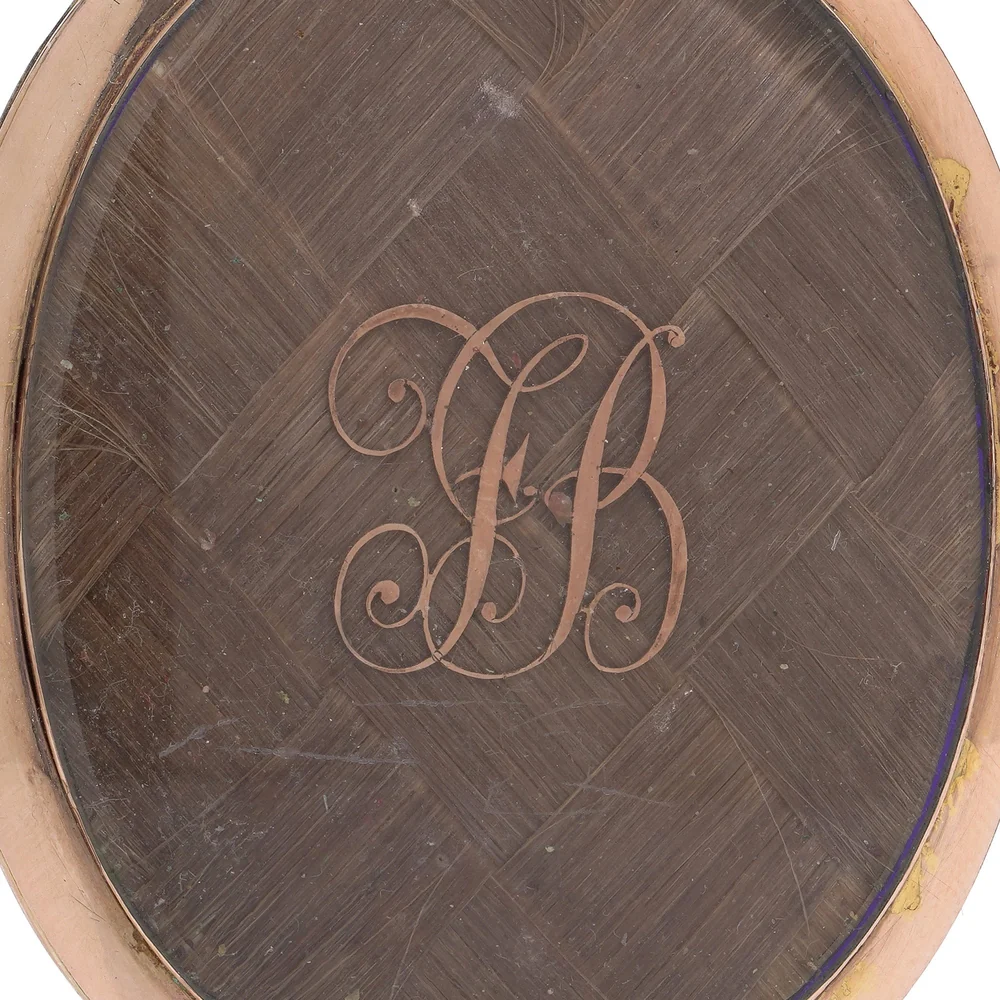 The reverse of a Georgian mourning pendant: brown hair woven in a basket lattice under glass, with a gold wire monogram FB laid on top