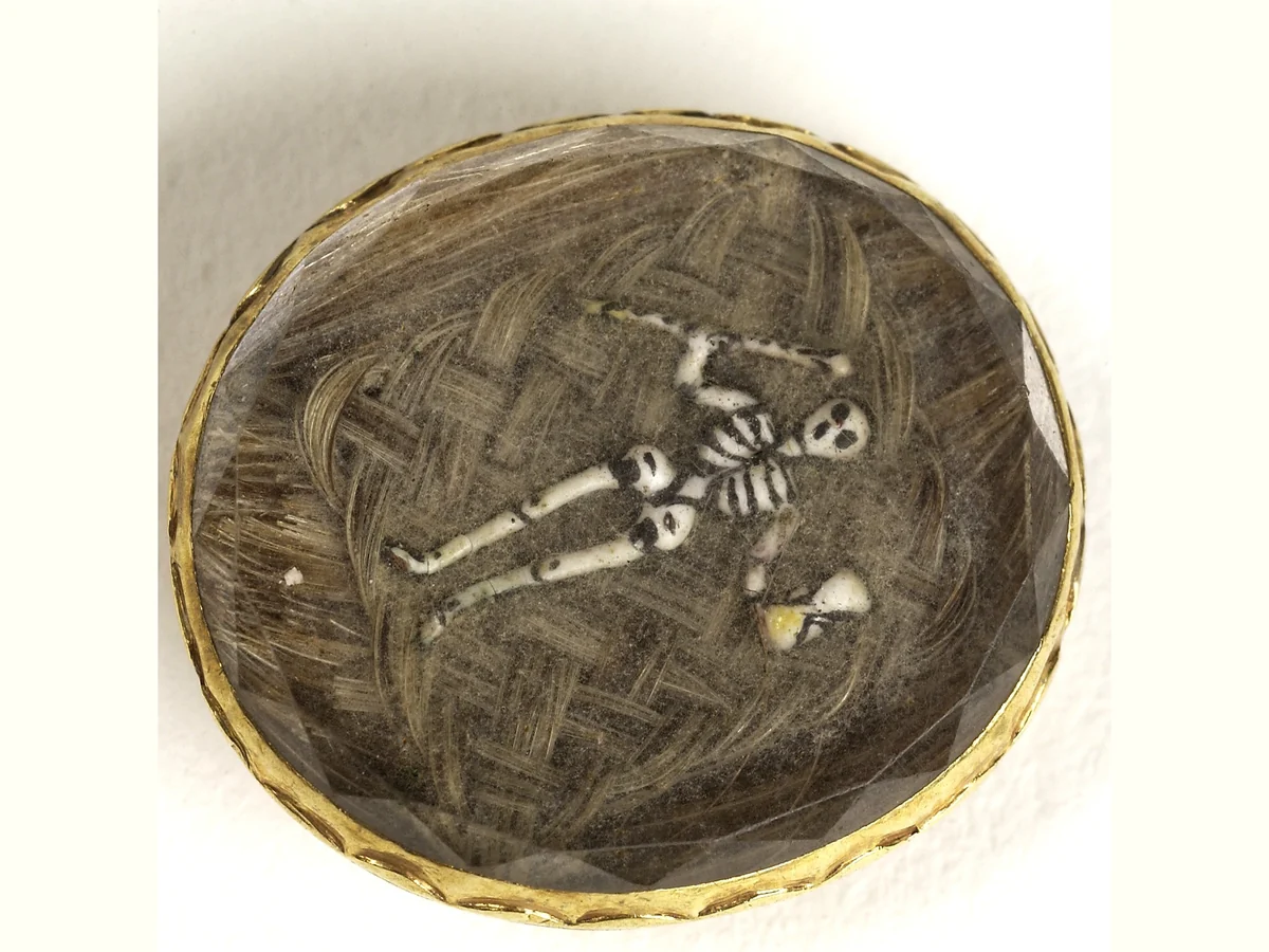A small oval mourning slide under crystal: a tiny white enamel skeleton laid over a ground of woven hair, in a thin gold frame