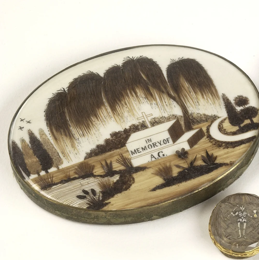 A group of mourning brooches: a large oval painted in sepia with weeping willows over a tomb inscribed In memory of, beside smaller brooches holding curls and plaits of hair, one framed in garnets and seed pearls