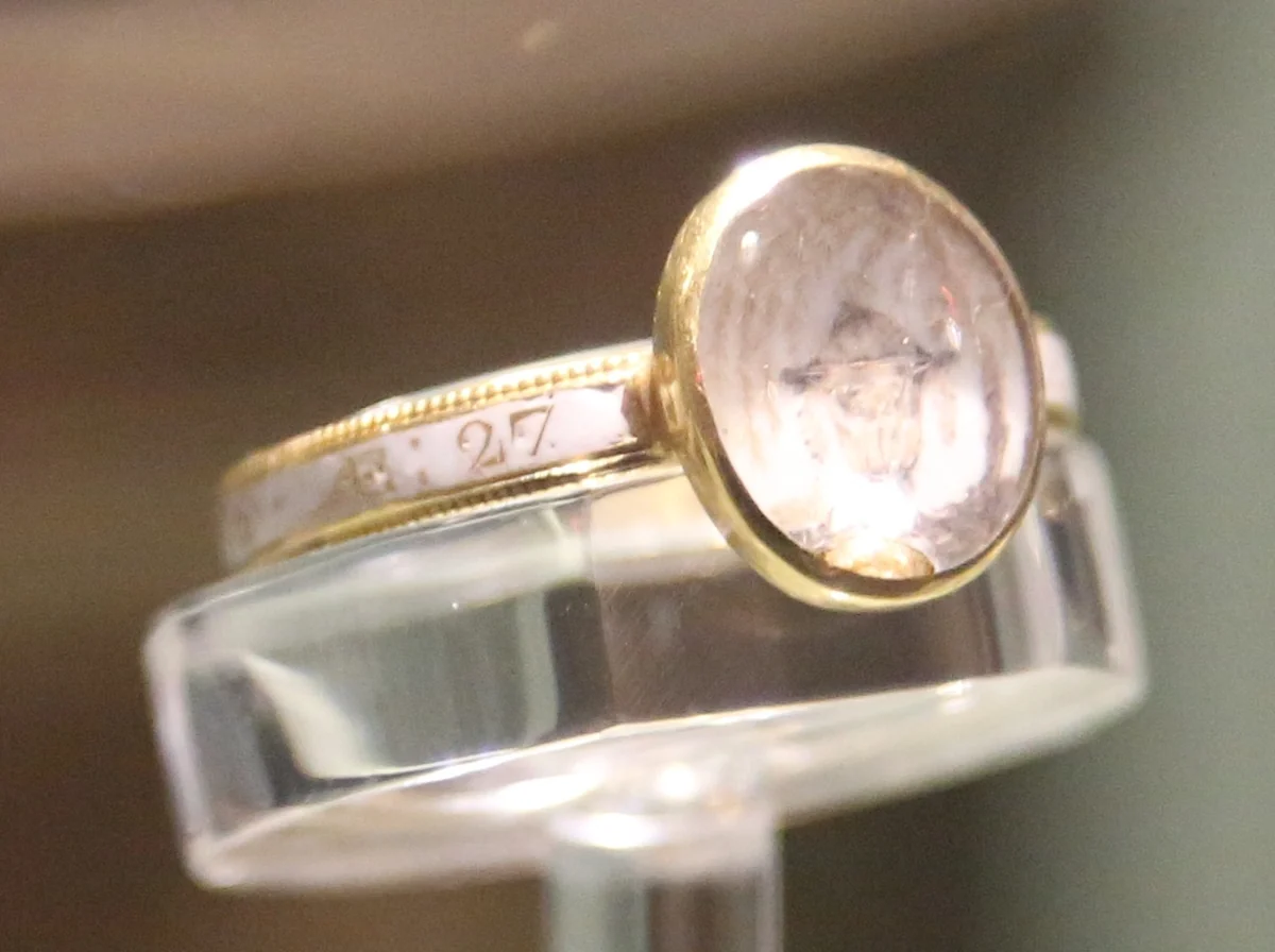 A gold mourning ring with a white enamel hoop and an oval crystal bezel, displayed on a clear acrylic stand