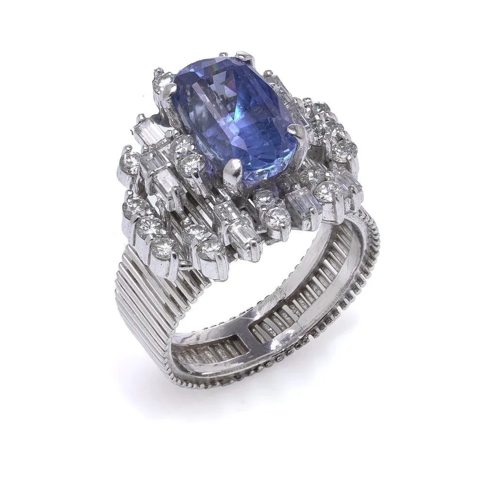 18KT white gold ring with a 3 ct oval Ceylon sapphire in a tiered cluster of baguette and round diamonds on a broad ribbed band