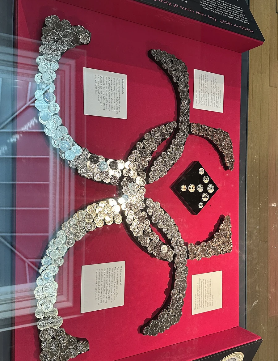 A British Museum display case of new King Charles III coins laid out in curved bands on a red background