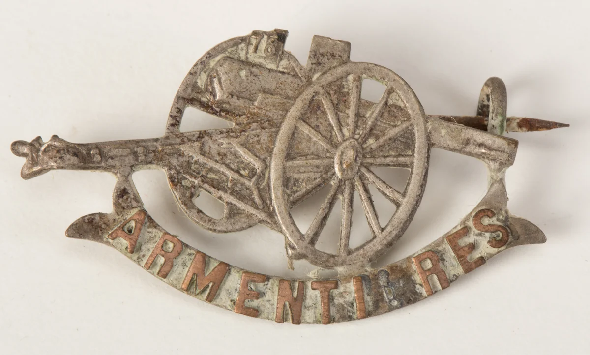A First World War place brooch: a silver field gun above a gold scroll reading ARMENTIERES, with the pin visible behind