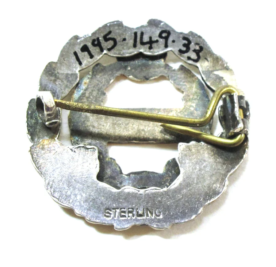 The back of a First World War silver sweetheart brooch: a hinged pin, a simple hook catch and the word STERLING stamped on the frame, with the museum number written in ink