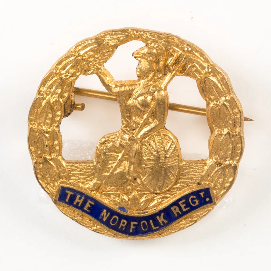 A gilt Norfolk Regiment sweetheart brooch showing seated Britannia inside a wreath, with a blue enamel scroll reading THE NORFOLK REGT.
