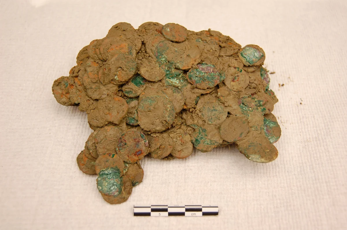 A lump of Roman coins from the Frome Hoard, stuck together with mud and green corrosion, beside a scale bar