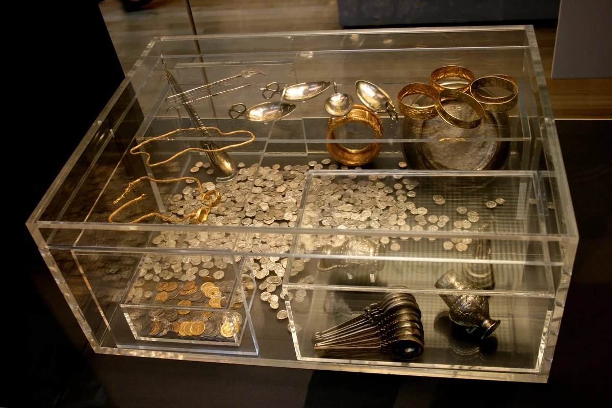 A clear display case showing the Hoxne Hoard laid out as it was excavated: piles of Roman silver and gold coins, gold bracelets, silver spoons and pepper pots
