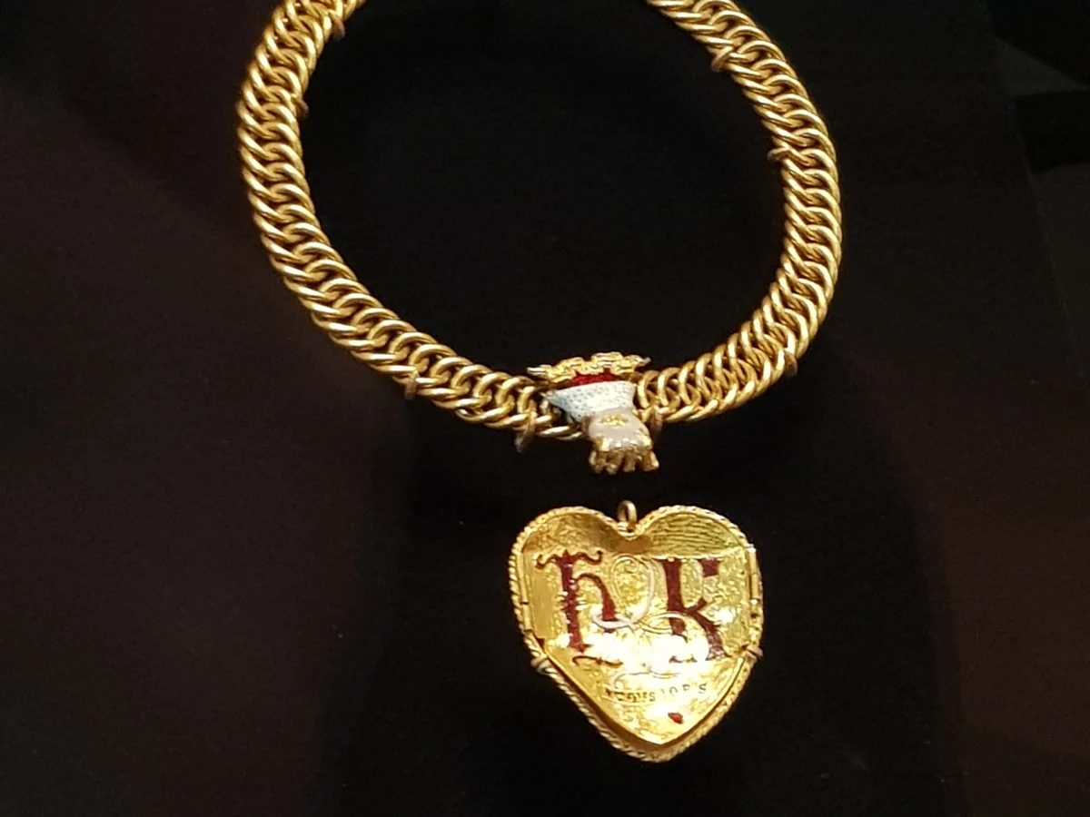 The Tudor Heart pendant: a gold heart with red enamelled initials H and K hanging from a woven gold chain, on a dark background