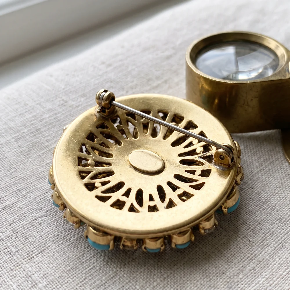 The back of a vintage gilt costume brooch with a pierced back plate, oval plaque, pin and rolling safety catch, beside a brass loupe