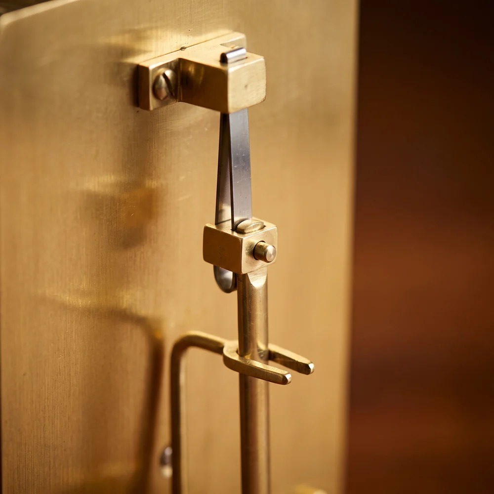 Close-up of a thin steel suspension spring, the top of the pendulum rod and the crutch fork behind a brass clock movement